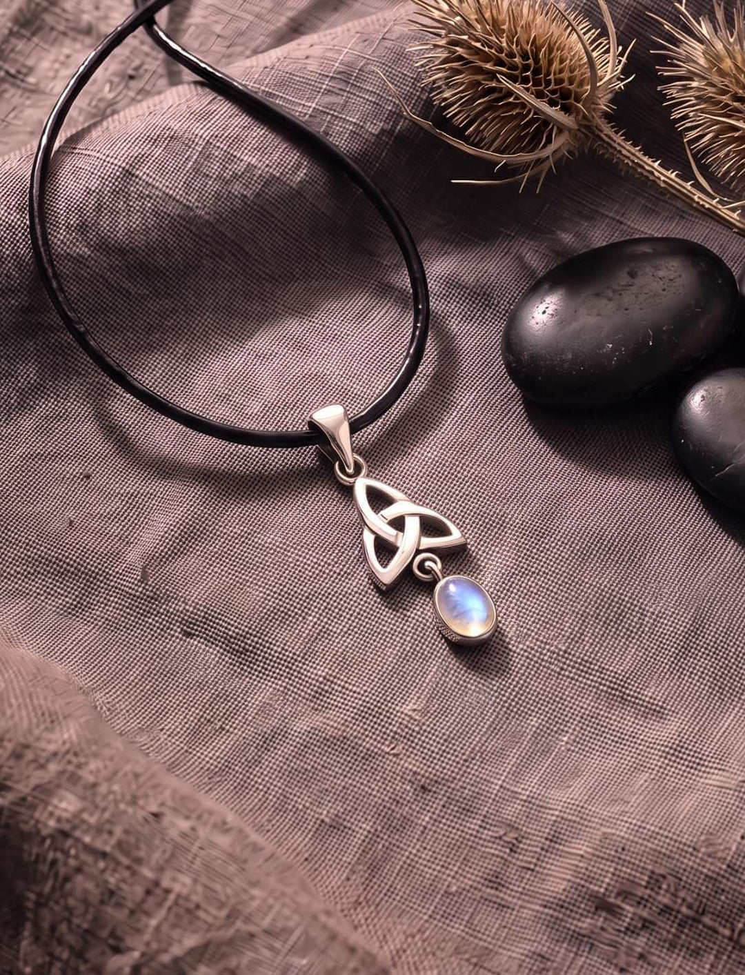 Triquetra Anhänger mit Regenbogenmondstein auf grauem Leinen neben getrocknetem Farn
