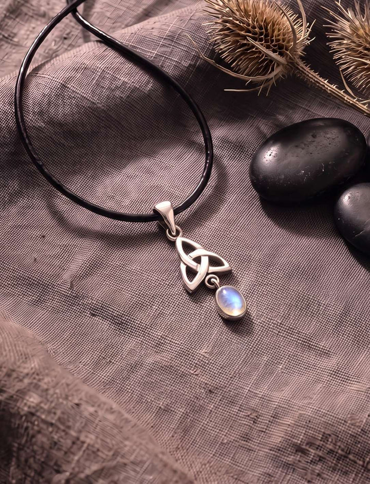 Triquetra Anhänger mit Regenbogenmondstein auf grauem Leinen neben getrocknetem Farn
