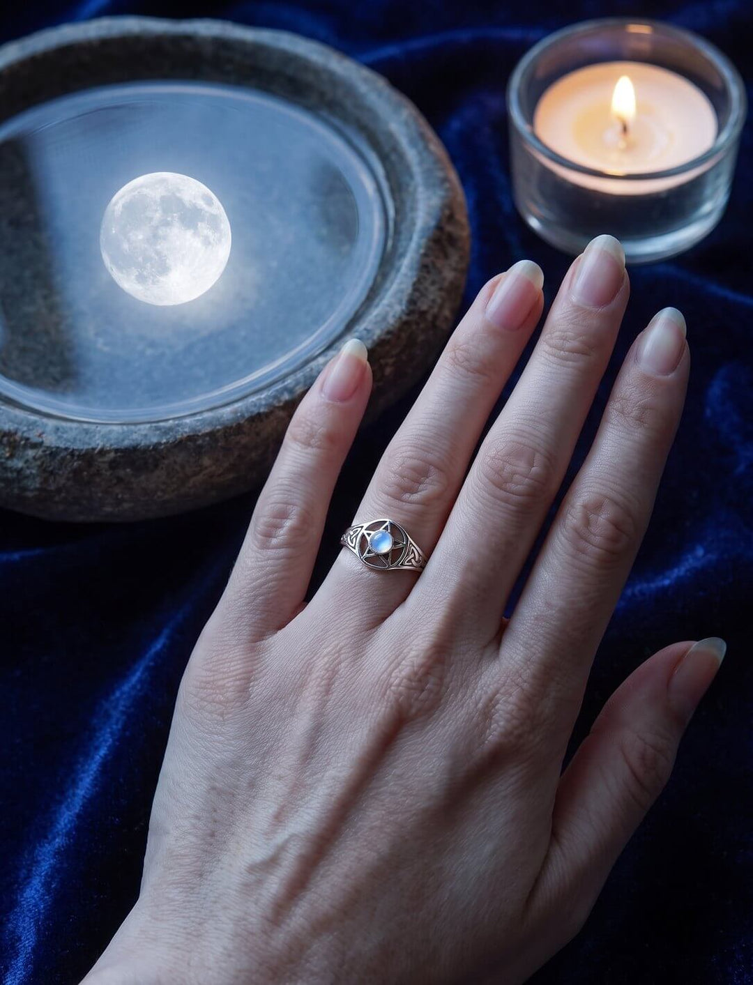 Pentakel Ring mit Regenbogenmondstein an der Hand auf blauem Samt, dahinter eine Wasserschale mit Vollmond