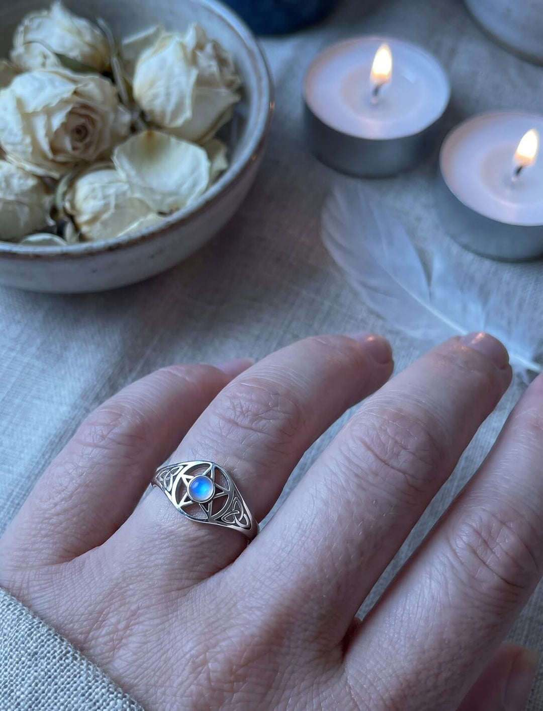 Drudenfuß-Ring mit blau schimmerndem Mondstein an der Hand, im Hintergrund weiße Rosenblüten und Teelichter