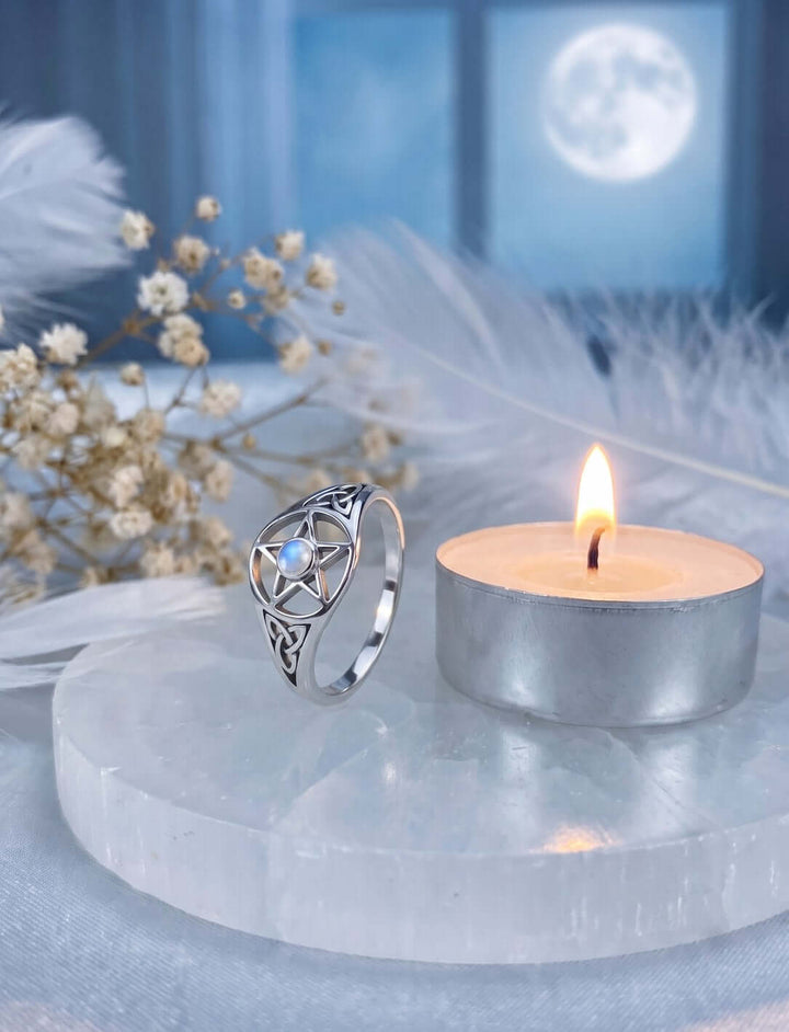 Mondstern der Triquetra: Pentagramm Ring aus Silber mit Regenbogenmondstein auf Selenit neben einem Teelicht, im Fenster der Vollmond