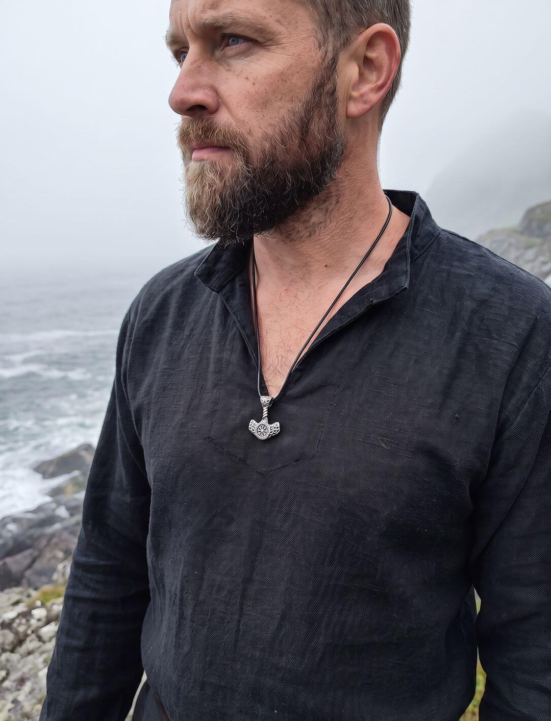 Mann im schwarzen Leinenhemd trägt einen Thors Hammer Anhänger an schwarzer Lederkordel an der nebligen Nordküste