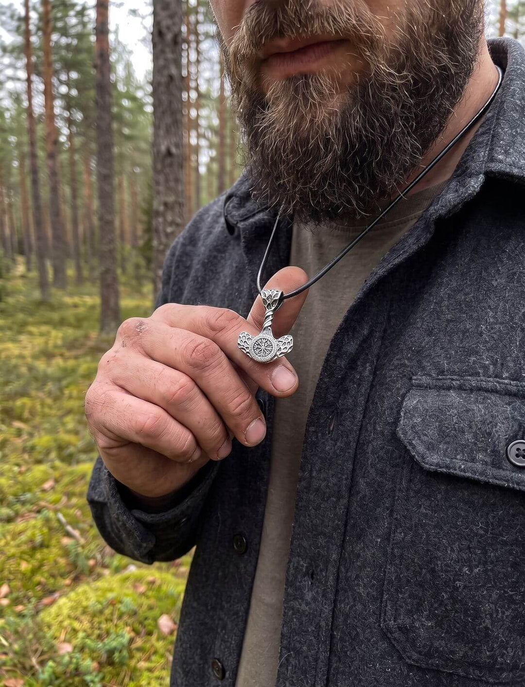 Bärtiger Mann im Wollhemd hält den kleinen Mjölnir-Anhänger an schwarzer Lederkordel zwischen den Fingern im Kiefernwald