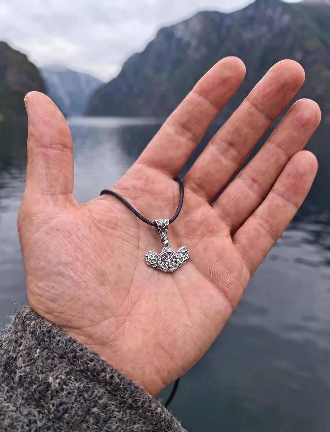 Größenvergleich: Thors Hammer mit Vegvísir, 3,1 x 2,3 cm, an schwarzer Lederkordel in einer offenen Männerhand am Fjord