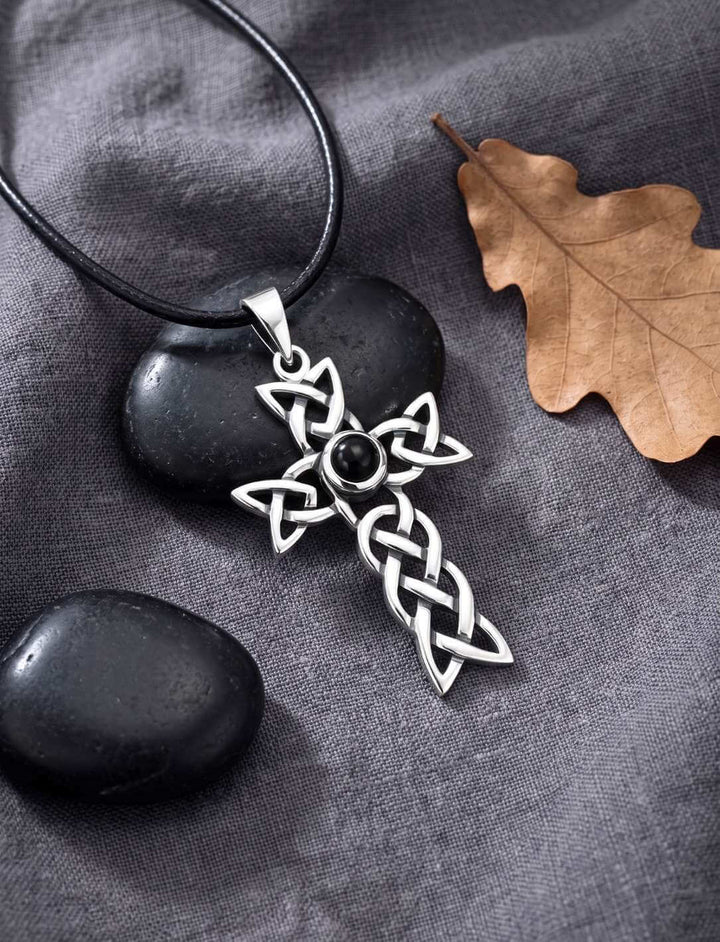 Silberkreuz mit schwarzem Onyx am Lederband auf grauem Leinen neben Kieseln und Eichenblatt