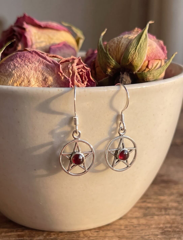 Glutstern: Pentagramm Ohrringe aus Silber mit Granat hängen an einer Keramikschale mit getrockneten Rosen