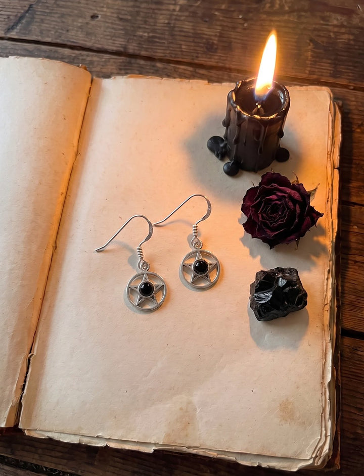 Pentagramm Ohrringe mit Onyx auf einem alten Buch neben schwarzer Kerze, getrockneter Rose und rohem Onyx