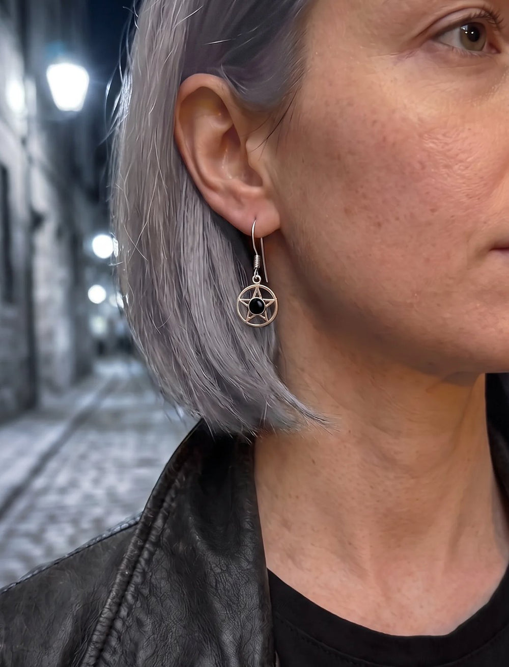 Frau mit grauem Bob und Lederjacke trägt Pentagramm Ohrringe mit Onyx in einer nächtlichen Gasse