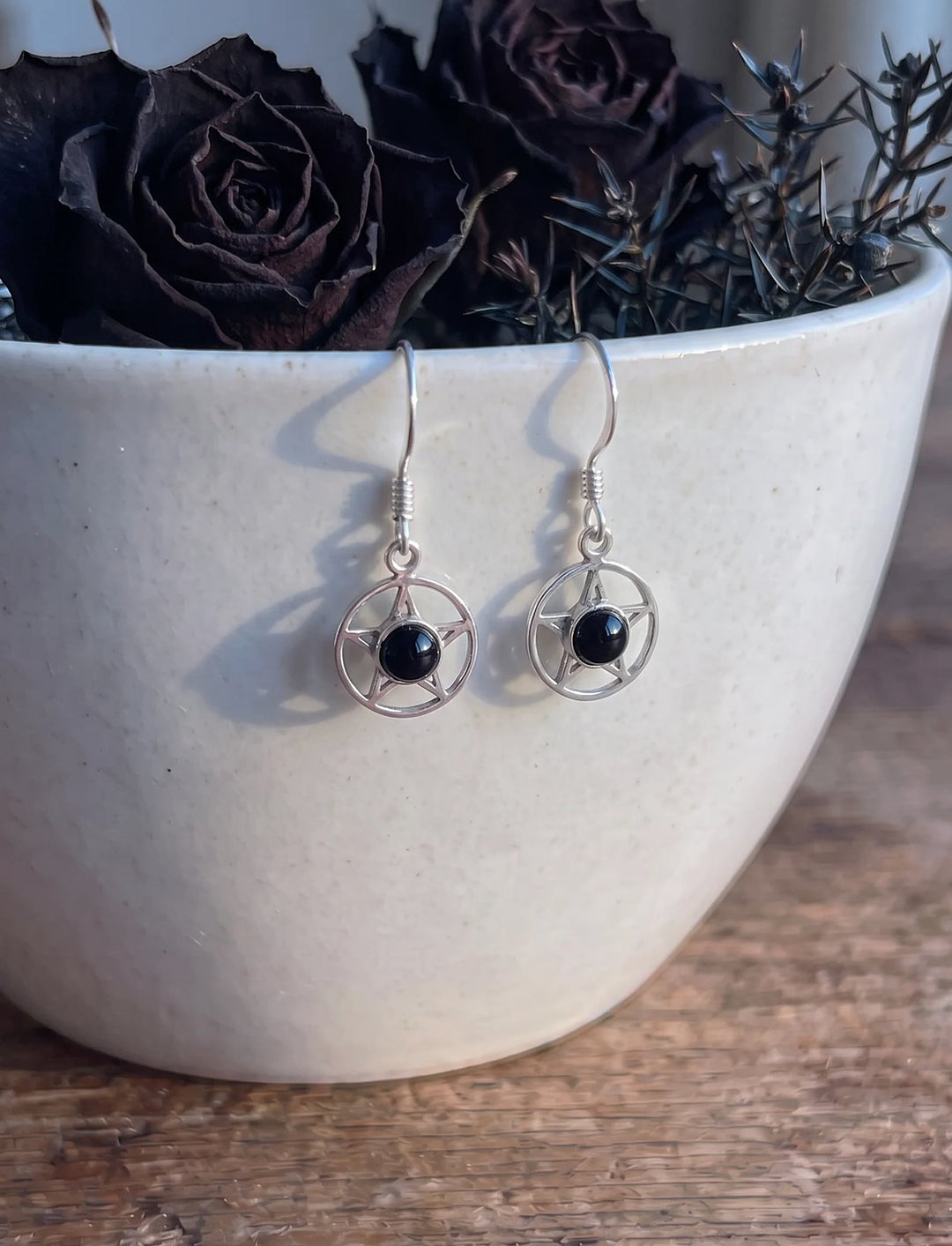 Pentagramm Ohrringe aus Silber mit schwarzem Onyx hängen am Rand einer hellen Keramikschale mit getrockneten schwarzen Rosen