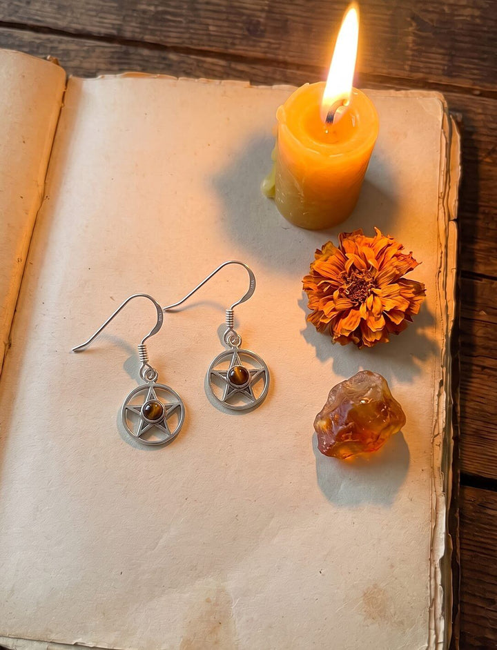 Ohrhänger aus 925 Silber mit Pentakel und Tigerauge auf einem alten Buch neben Bienenwachskerze, Ringelblume und Bernstein