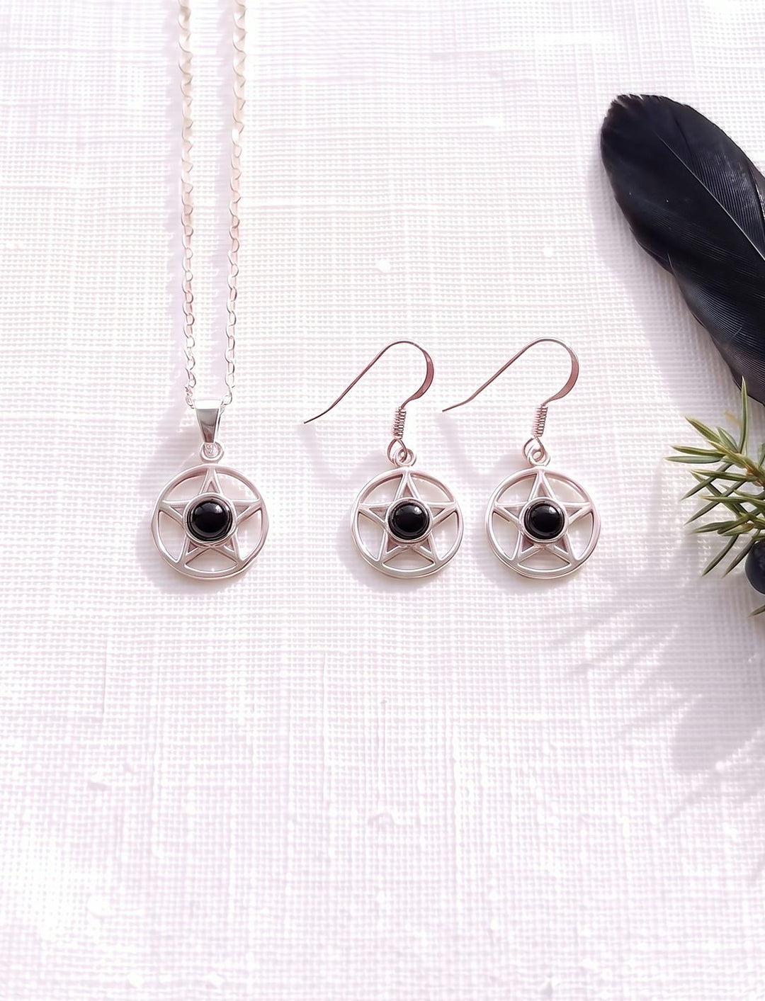 Schmuckset aus Pentagramm Kette und Ohrringen mit Onyx in Silber auf weißem Leinen neben Rabenfeder und Wacholder