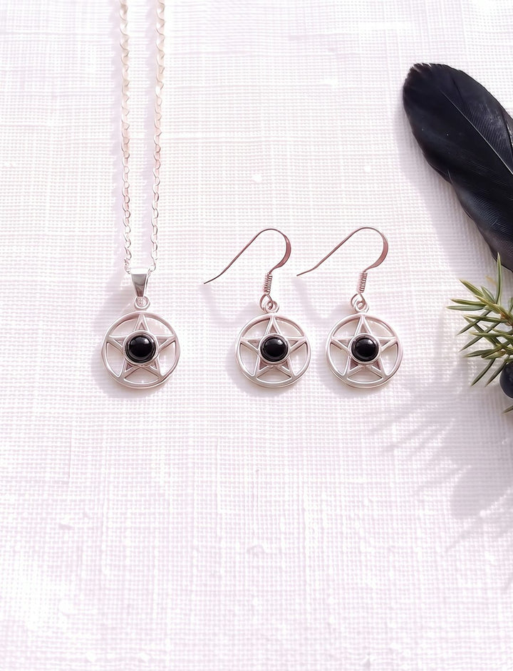 Schmuckset aus Pentagramm Kette und Ohrringen mit Onyx in Silber auf weißem Leinen neben Rabenfeder und Wacholder