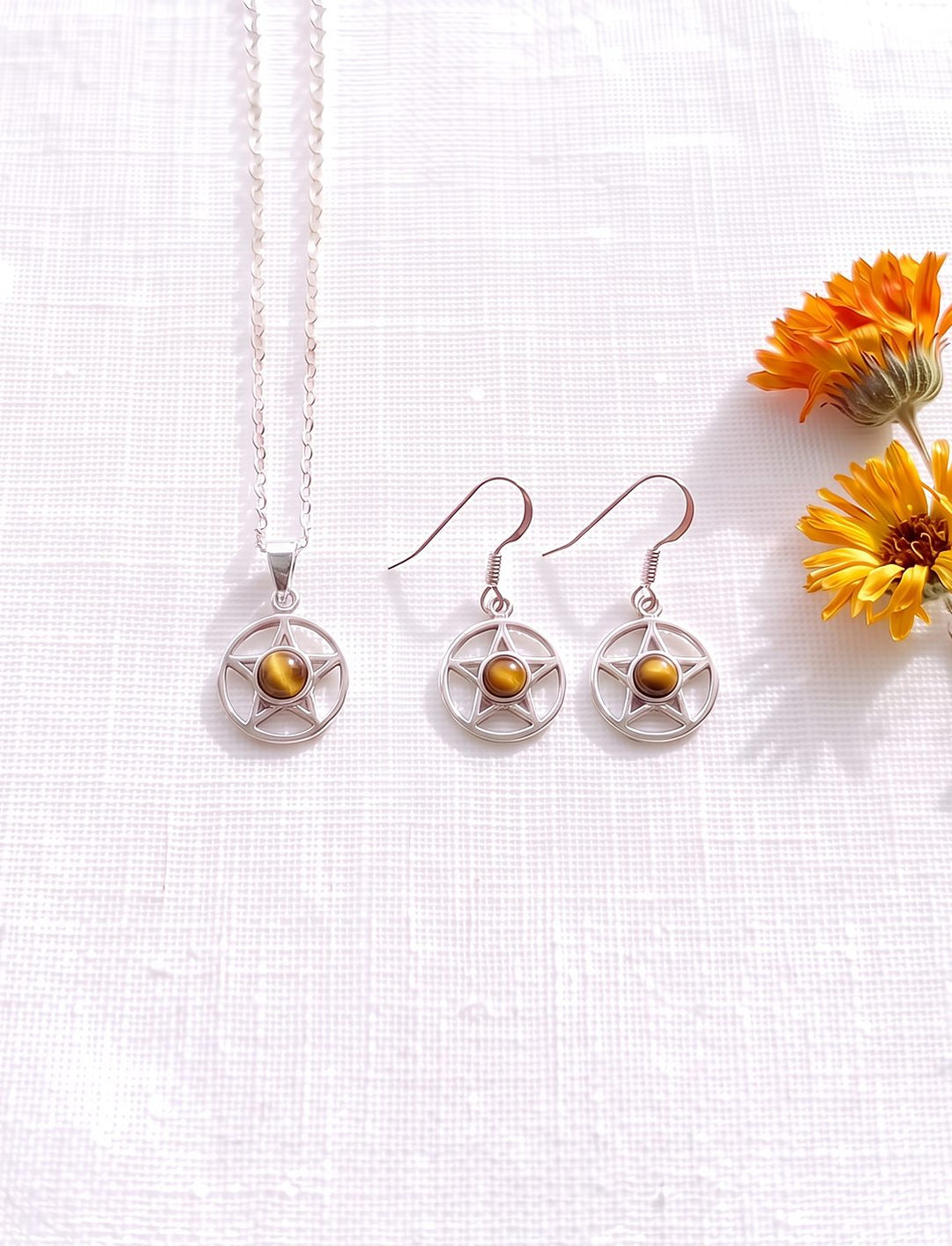 Schmuckset aus Pentagramm Kette und Ohrringen mit Tigerauge in Silber auf weißem Leinen neben Ringelblumen