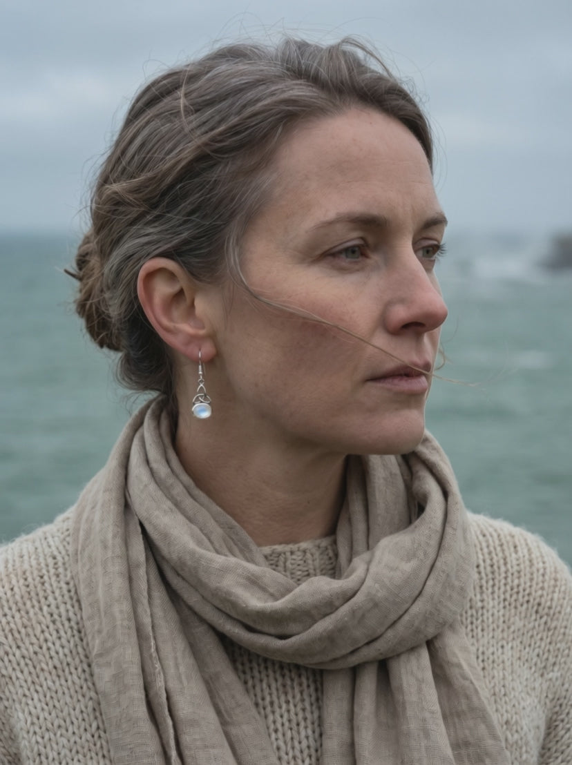 Frau mit Triquetra Ohrhänger aus Silber und Mondstein blickt an der irischen Küste aufs Meer, Wind im Haar
