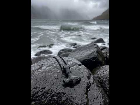 Video: Schwarzer Thors Hammer Anhänger liegt auf nassem Fels, dahinter rollen Wellen in einem nebligen Fjord