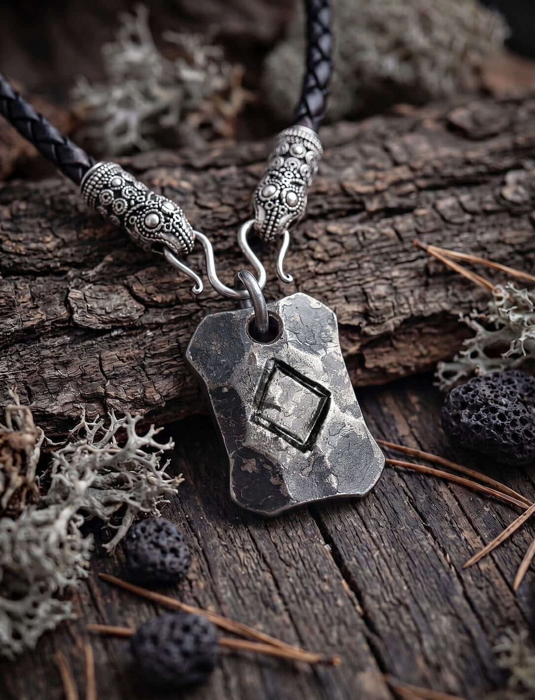 Ingwaz Rune Anhänger an geflochtener Lederkette mit versilberten Rabenköpfen auf Rinde mit Flechten und Lavasteinen