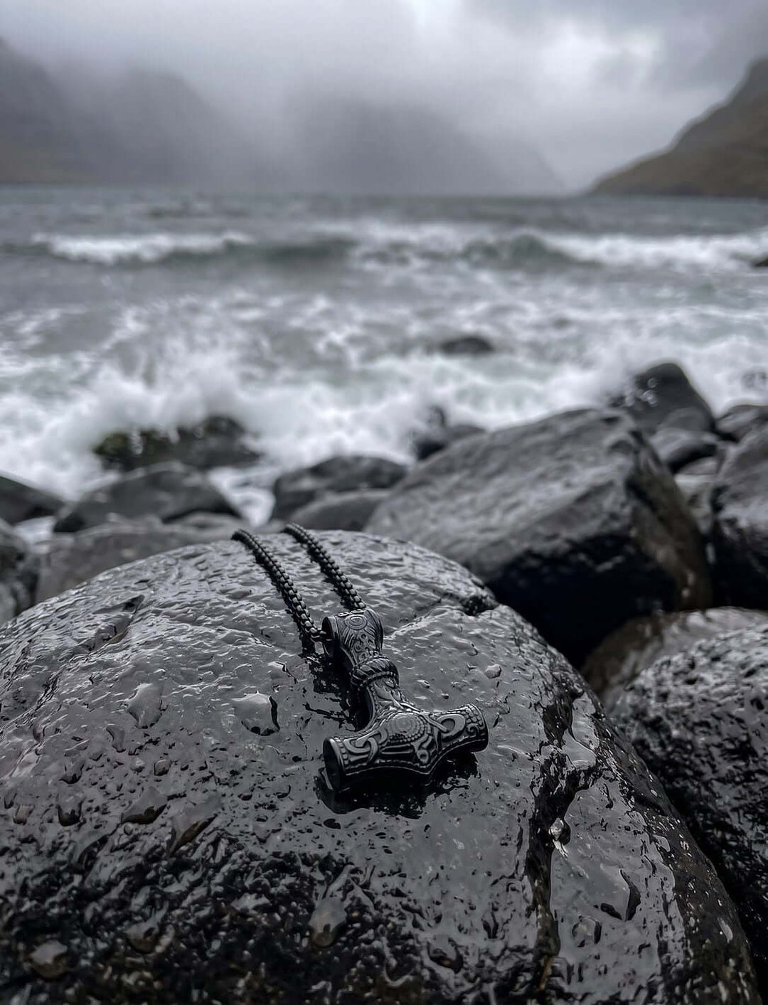 Thors Hammer Kette in Schwarz auf nassem Basaltfels an einer stürmischen Fjordküste im Nebel