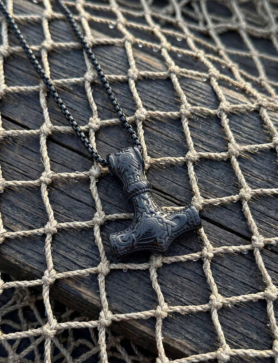 Schwarzer Thors Hammer aus Edelstahl liegt auf einem alten Fischernetz über dunklem Bootsholz
