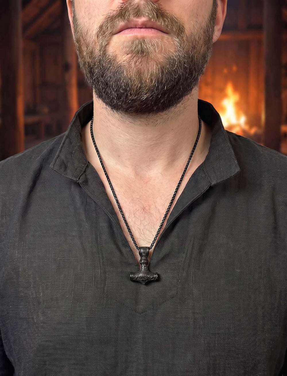 Mann im dunklen Leinenhemd mit schwarzem Mjölnir-Anhänger auf der Brust, im Hintergrund Feuer in einem Langhaus