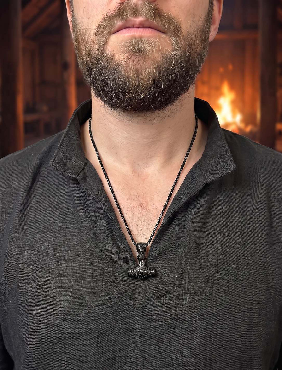 Mann im dunklen Leinenhemd mit schwarzem Mjölnir-Anhänger auf der Brust, im Hintergrund Feuer in einem Langhaus