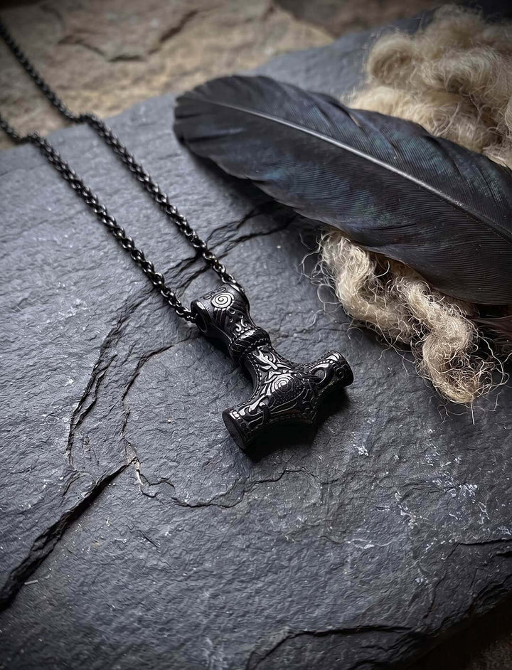 Schwarzer Donner: Thors Hammer Anhänger aus schwarzem Edelstahl an Kette auf Schiefer neben einer Rabenfeder