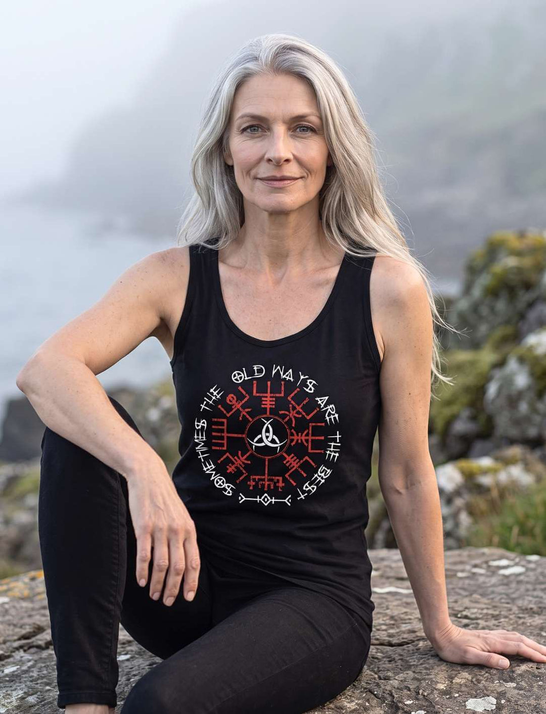Frau sitzt auf einem Küstenfelsen, Nahaufnahme des Vegvisir Motivs auf schwarzem Tank Top