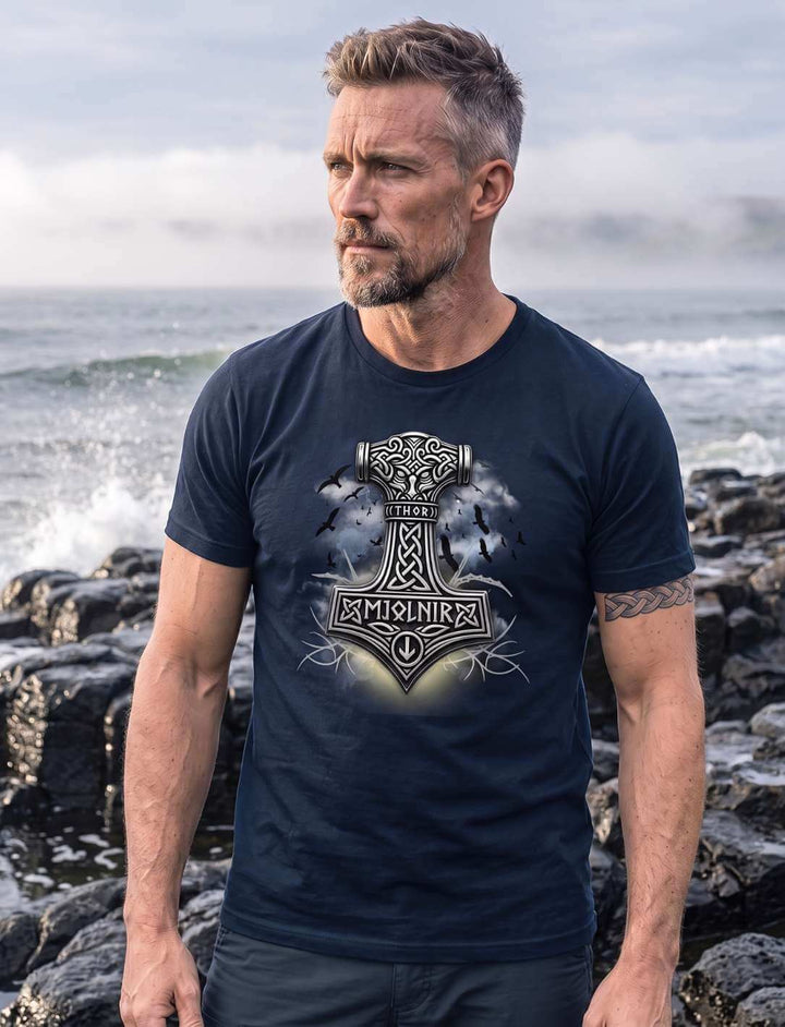 Mann in French Navy T-Shirt an der Basaltküste, Gischt hinter ihm, Thors Hammer mit Knotenwerk auf der Brust