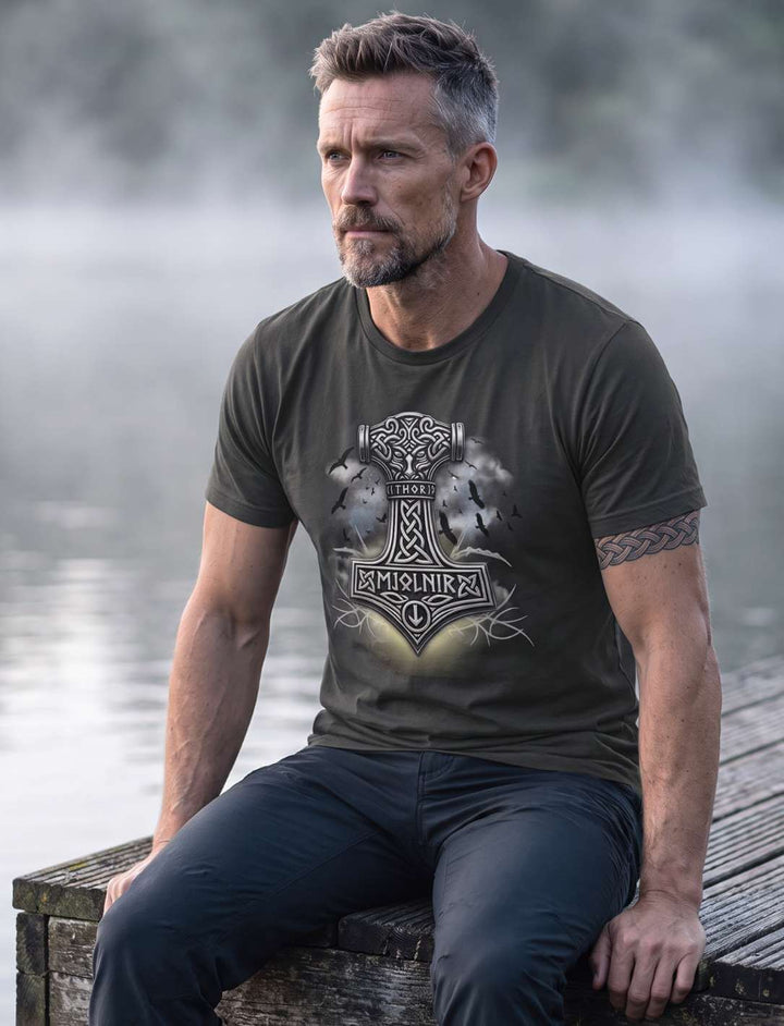 Mann in anthrazitfarbenem T-Shirt sitzt auf der Stegkante am See, Hammer-Motiv mit Rabenschwarm sichtbar