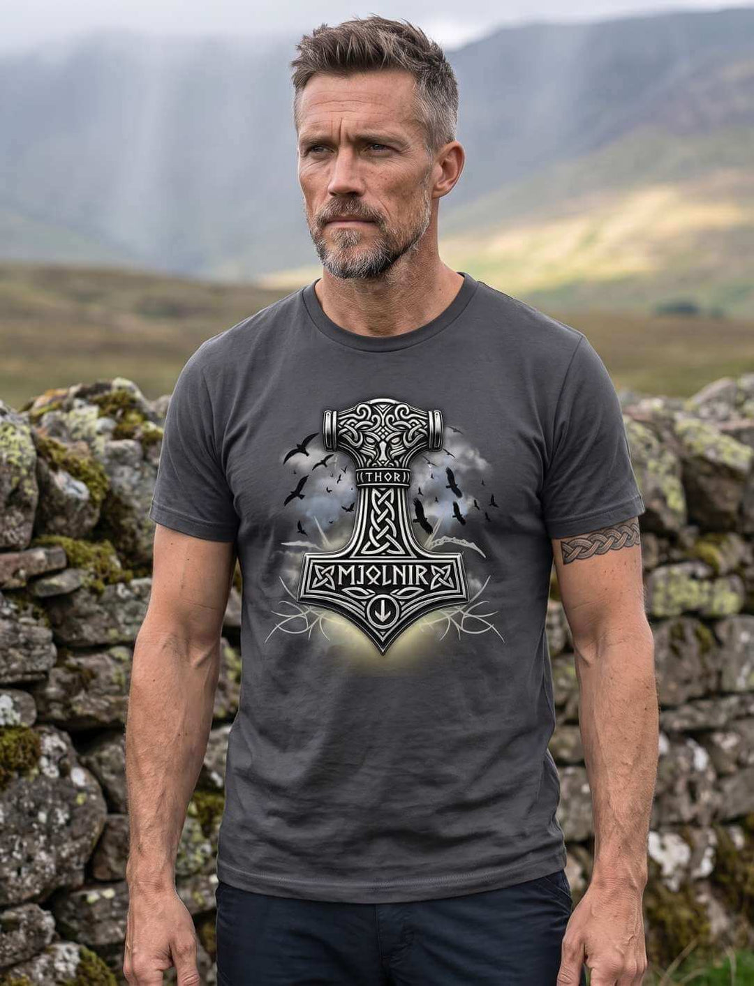 Mann in Anthrazit-Shirt vor moosiger Steinmauer auf der Hochebene, silbergrauer Hammer mit Schriftzug Thor