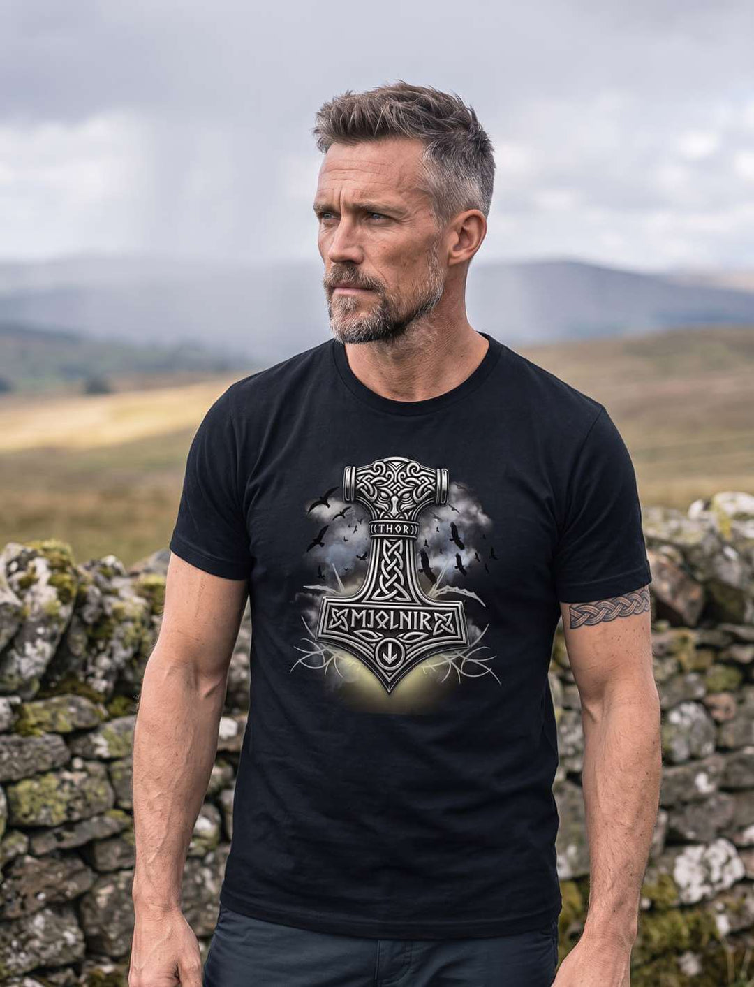 Mann in schwarzem T-Shirt steht an einer Trockensteinmauer, Wolkenschatten über der Heide, Hammer-Motiv gut lesbar