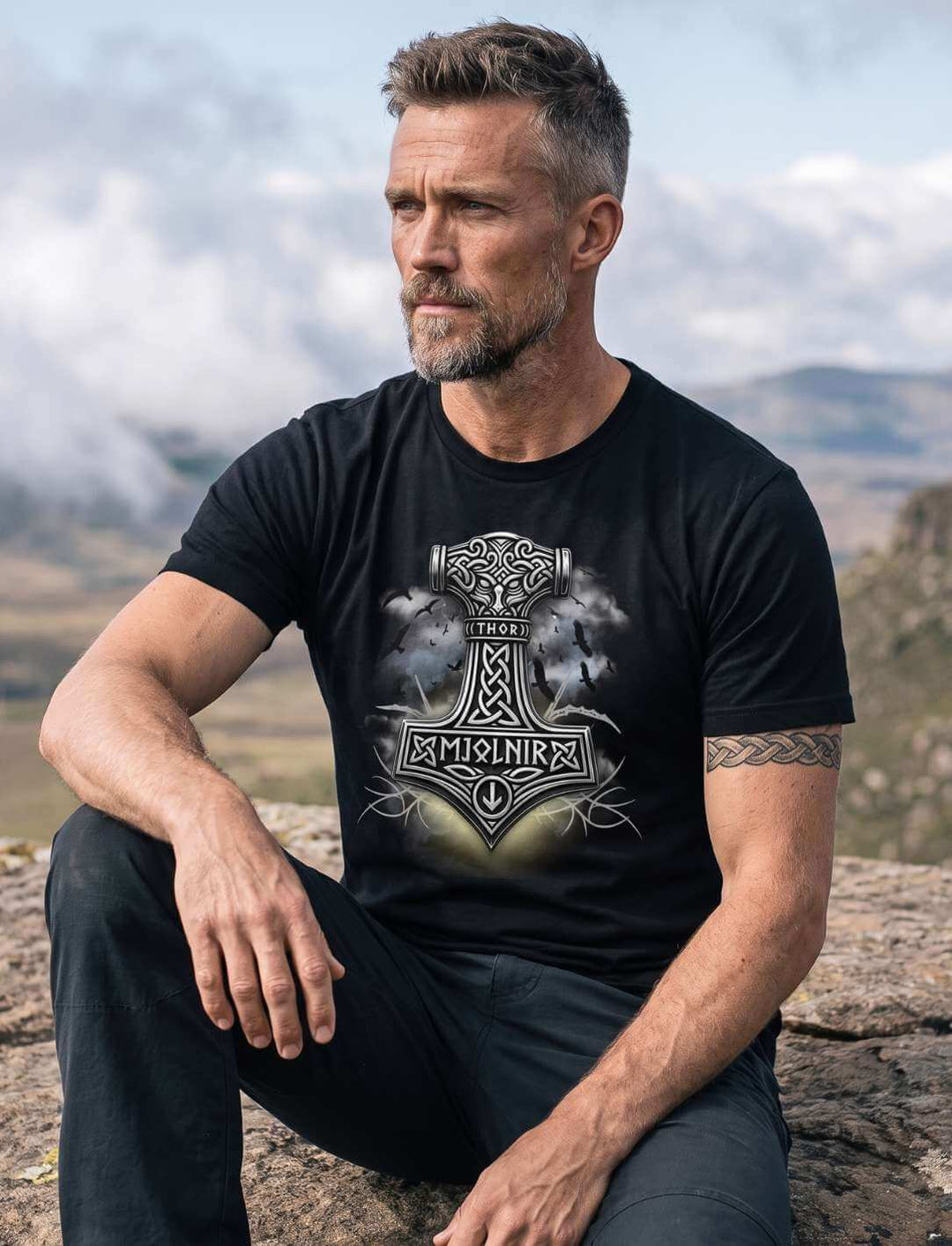 Mann in schwarzem T-Shirt sitzt auf einem Felsen vor weiter Hügellandschaft, Unterarme auf den Knien