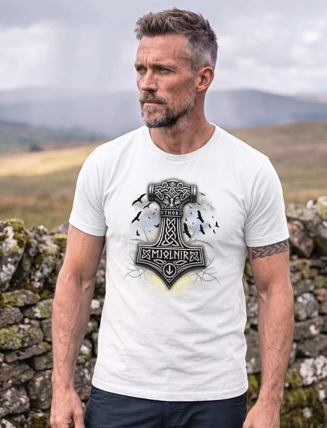 Mann in weißem T-Shirt mit Mjolnir-Druck vor einer Trockensteinmauer, Regenschauer über den Hügeln