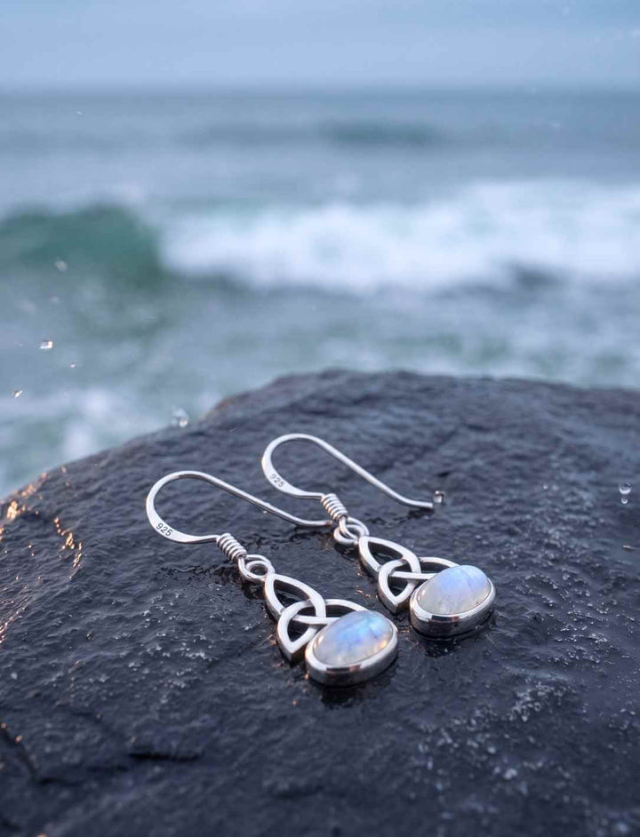 Triquetra Ohrringe aus Silber mit Mondstein auf nassem Küstenfels, Welle im Hintergrund