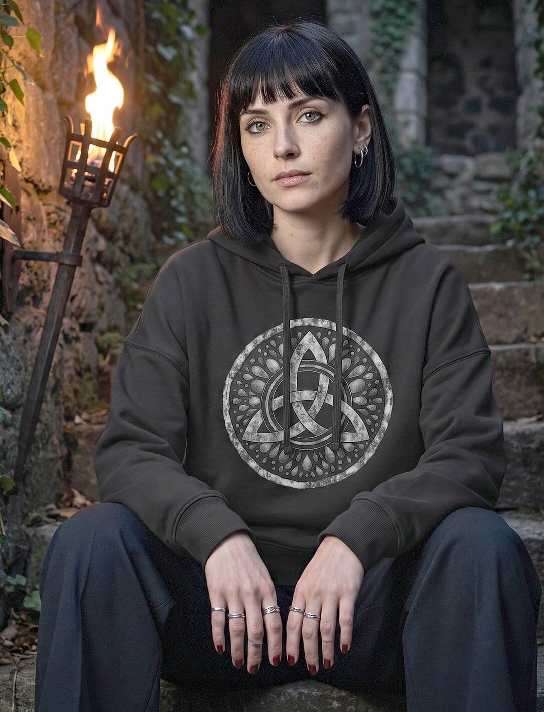 Frau im schwarzen Triquetra Boxcut Hoodie sitzt auf alten Steinstufen im Fackelschein