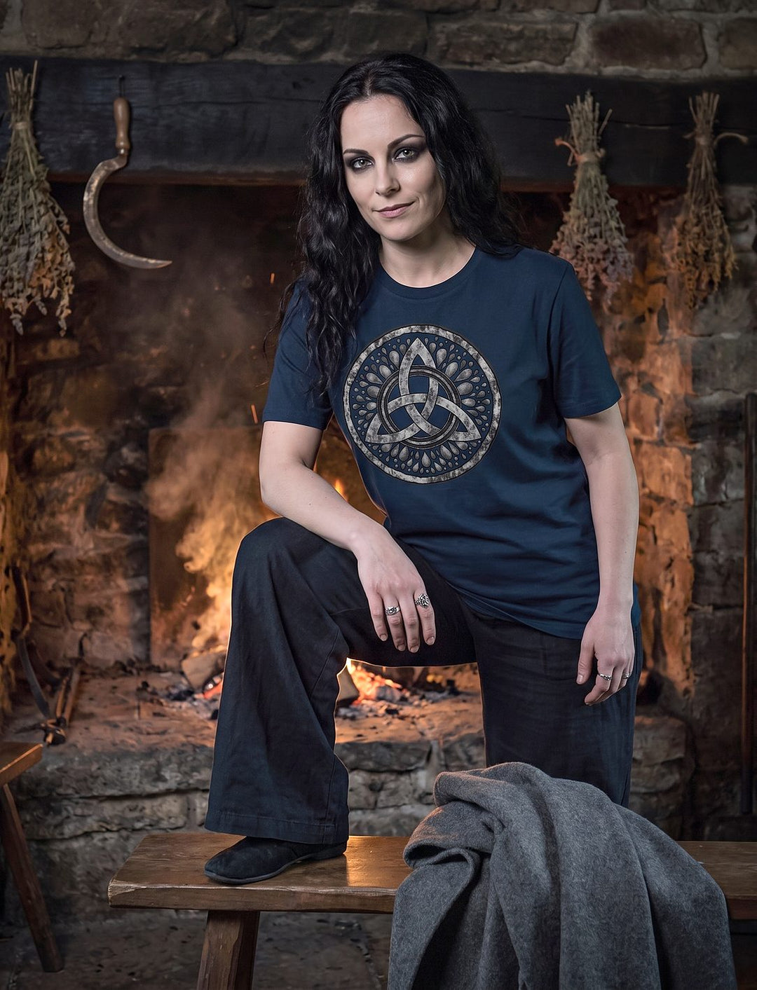 Frau im French Navy Damen Shirt mit Triquetra stützt den Fuß auf eine Bank vor dem Herdfeuer einer Steinhütte