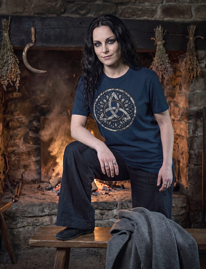 Frau im French Navy Damen Shirt mit Triquetra stützt den Fuß auf eine Bank vor dem Herdfeuer einer Steinhütte