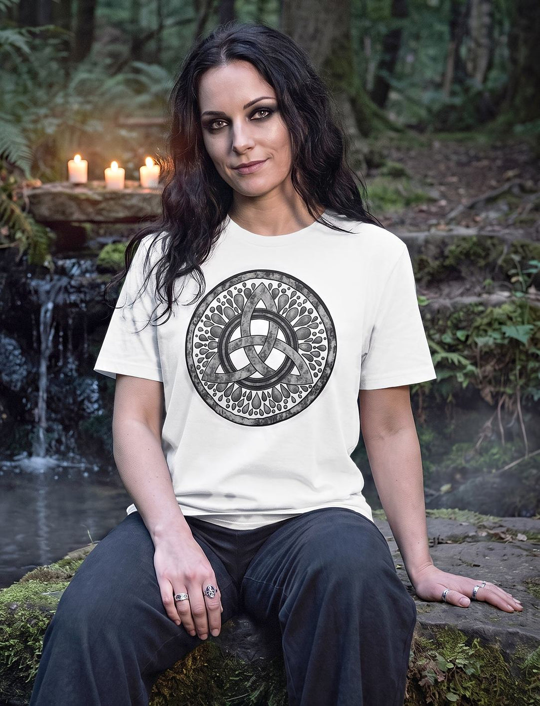 Frau im weißen Triquetra Shirt sitzt am Wasserfall einer Waldquelle, Kerzen leuchten im Hintergrund