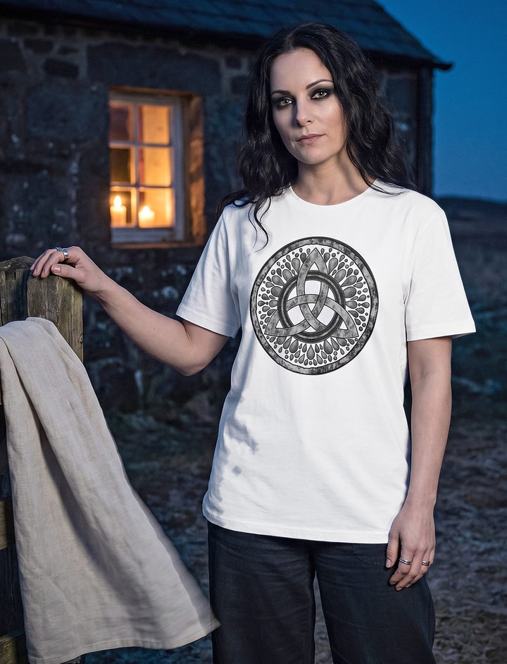 Frau im weißen Damen Shirt mit keltischem Knoten am Zaun vor einer Steinhütte mit Kerzenlicht im Fenster
