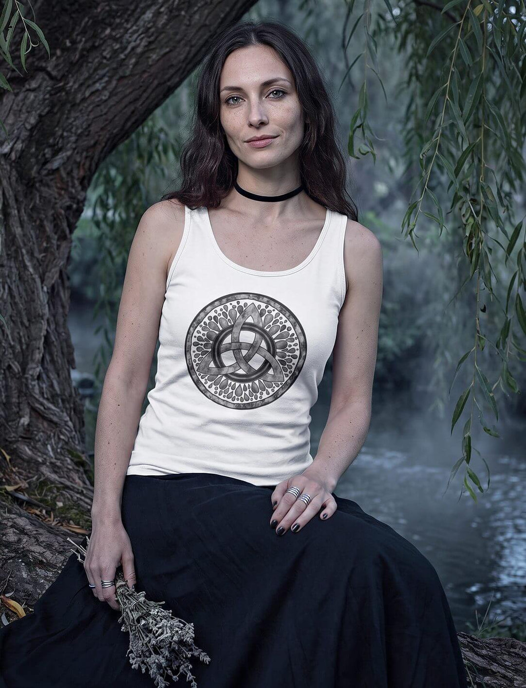 Frau im weißen Triquetra Tank Top sitzt unter einer Weide am Fluss, in der Hand ein Strauß Trockenblumen